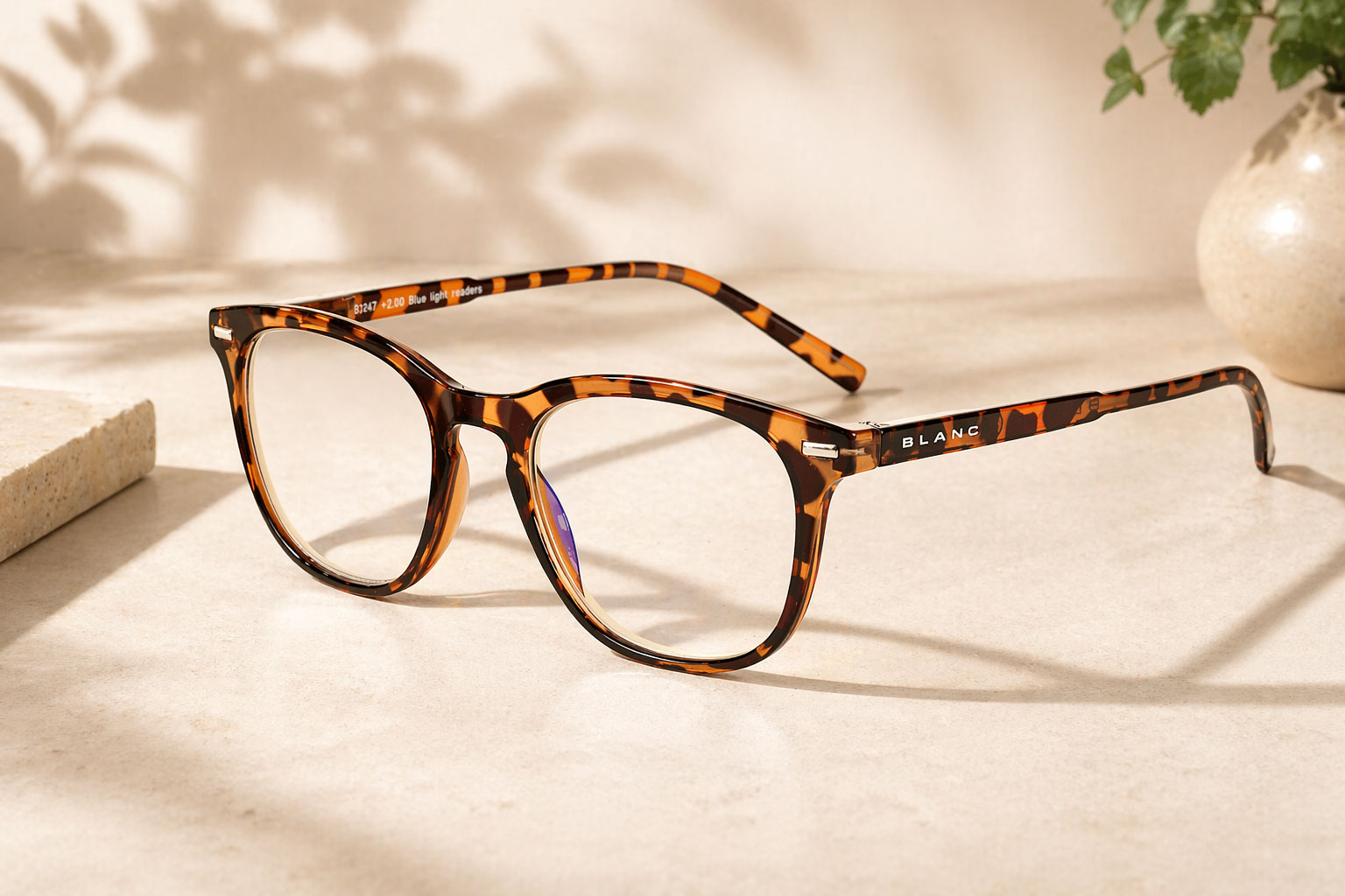 Ett par blåljusglasögon med runda linser vilar på en beige yta bredvid en stenplatta och en vas, med växtskuggor synliga i bakgrunden.