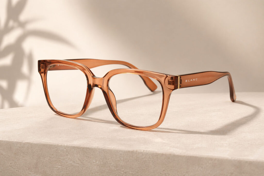 Ett par genomskinligt bruna läsglasögon med tjocka fyrkantiga bågar vilar på en ljusbeige stenyta. Ordet "BLANC" är tryckt i vitt på skalmen. Bakgrunden är neutral med en mjuk skugga.