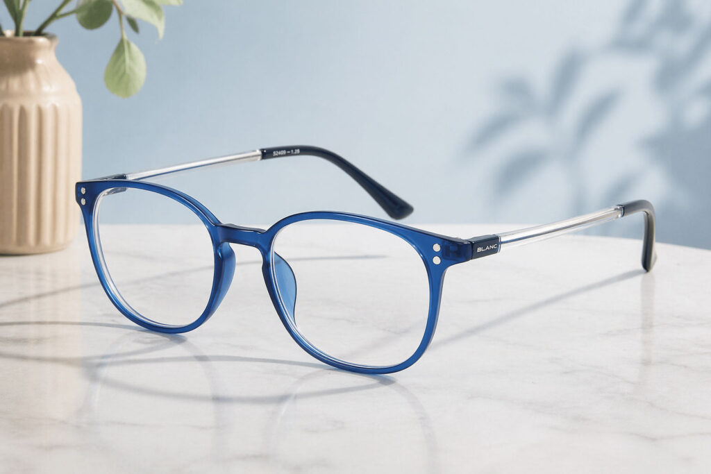 Ett par blåbågade minusglasögon med klara bågar och svarta spetsar vilar på en vit marmoryta; en beige vas med gröna blad och växtskuggor finns i bakgrunden.